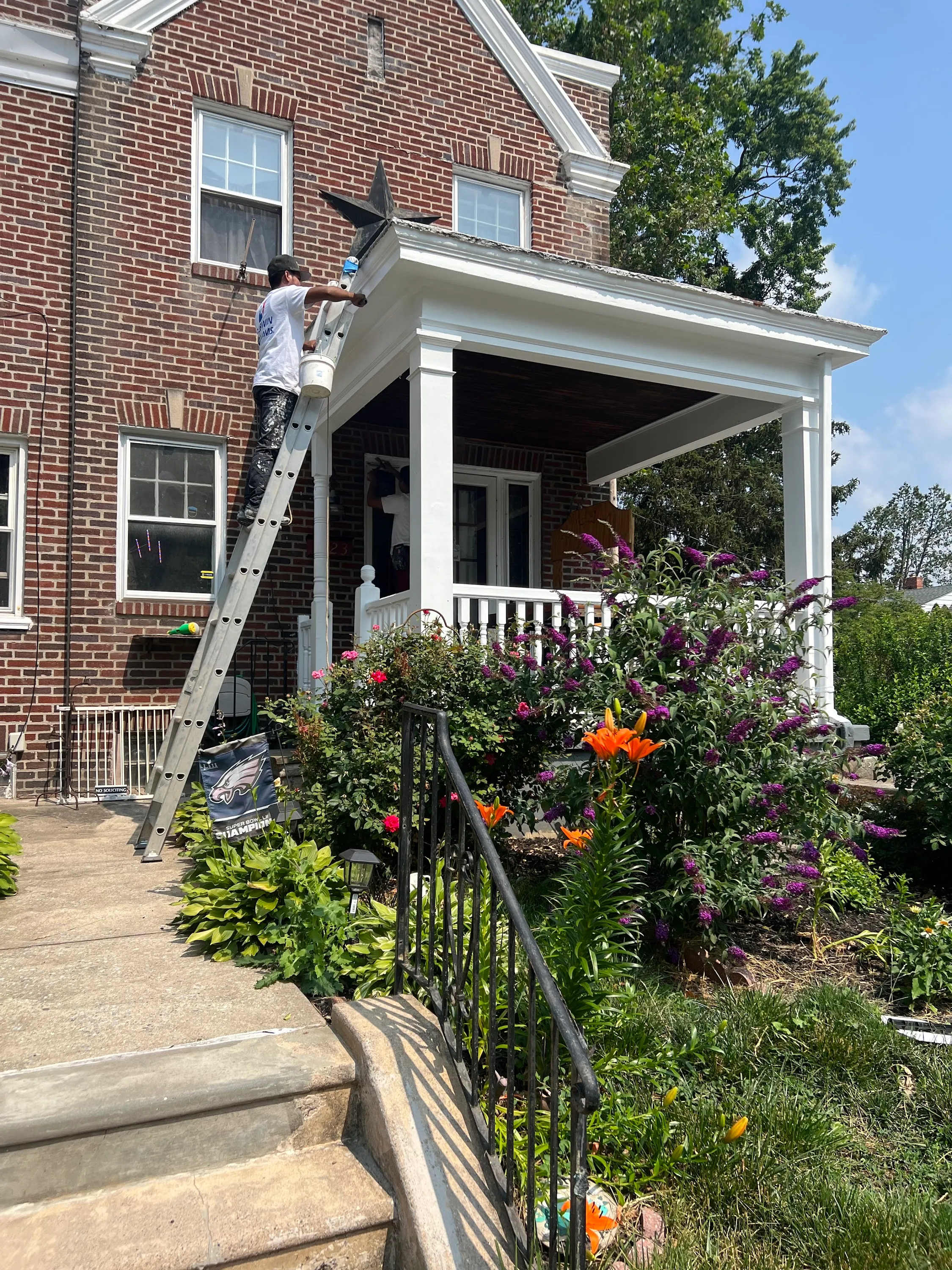 Red-brick Pennsylvania row house with white-painted front porch and JL crew on a tall extension ladder painting upper cornice trim