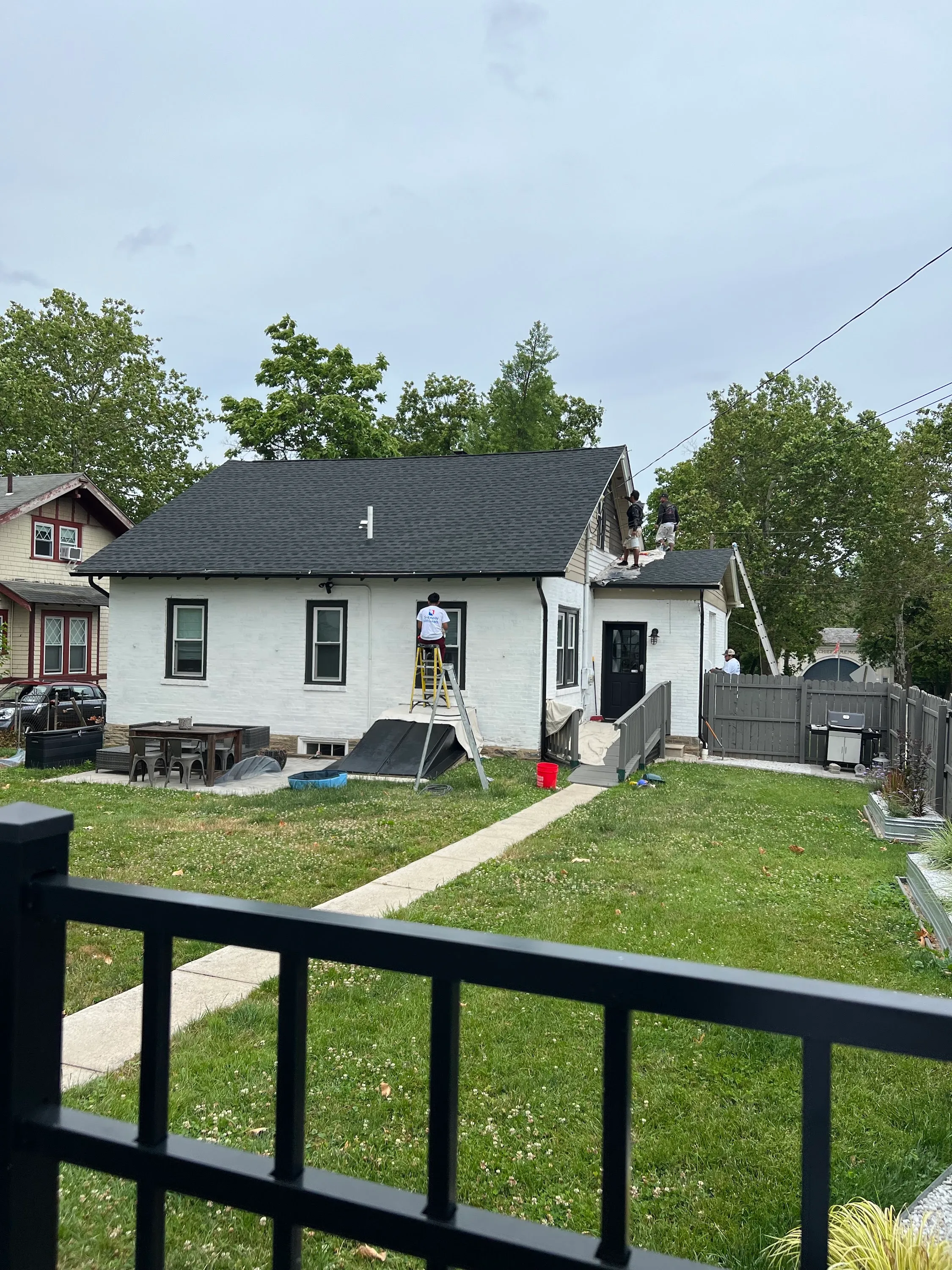 Painted-brick rear of a Pennsylvania home with a vinyl-sided shed dormer, JL crew member on a yellow ladder painting the upper trim, second crew member working the dormer roof