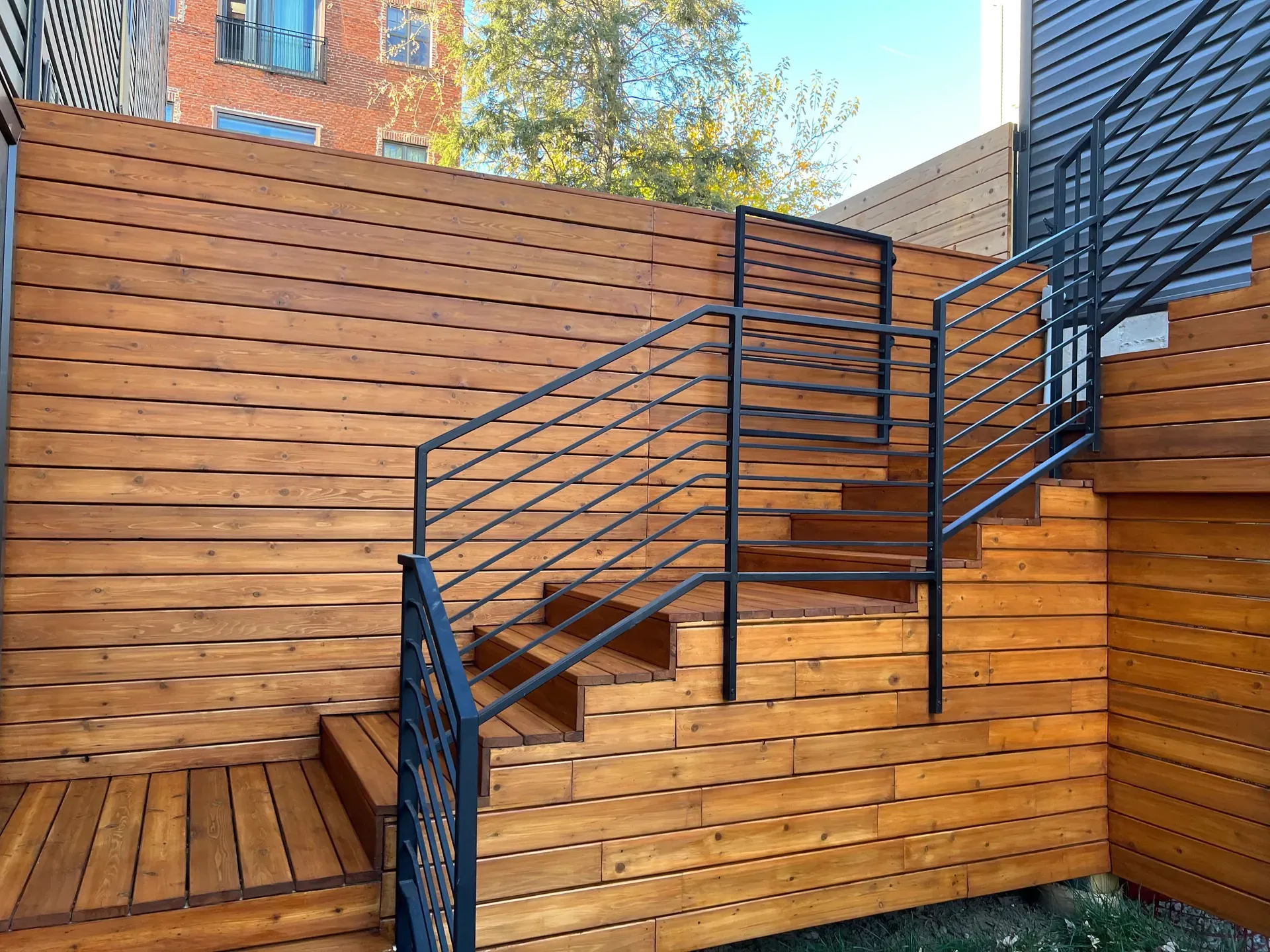 Finished cedar tongue-and-groove exterior stair with welded black steel railings, cedar siding wrapping the surround walls, urban rowhouse setting in late afternoon sunlight