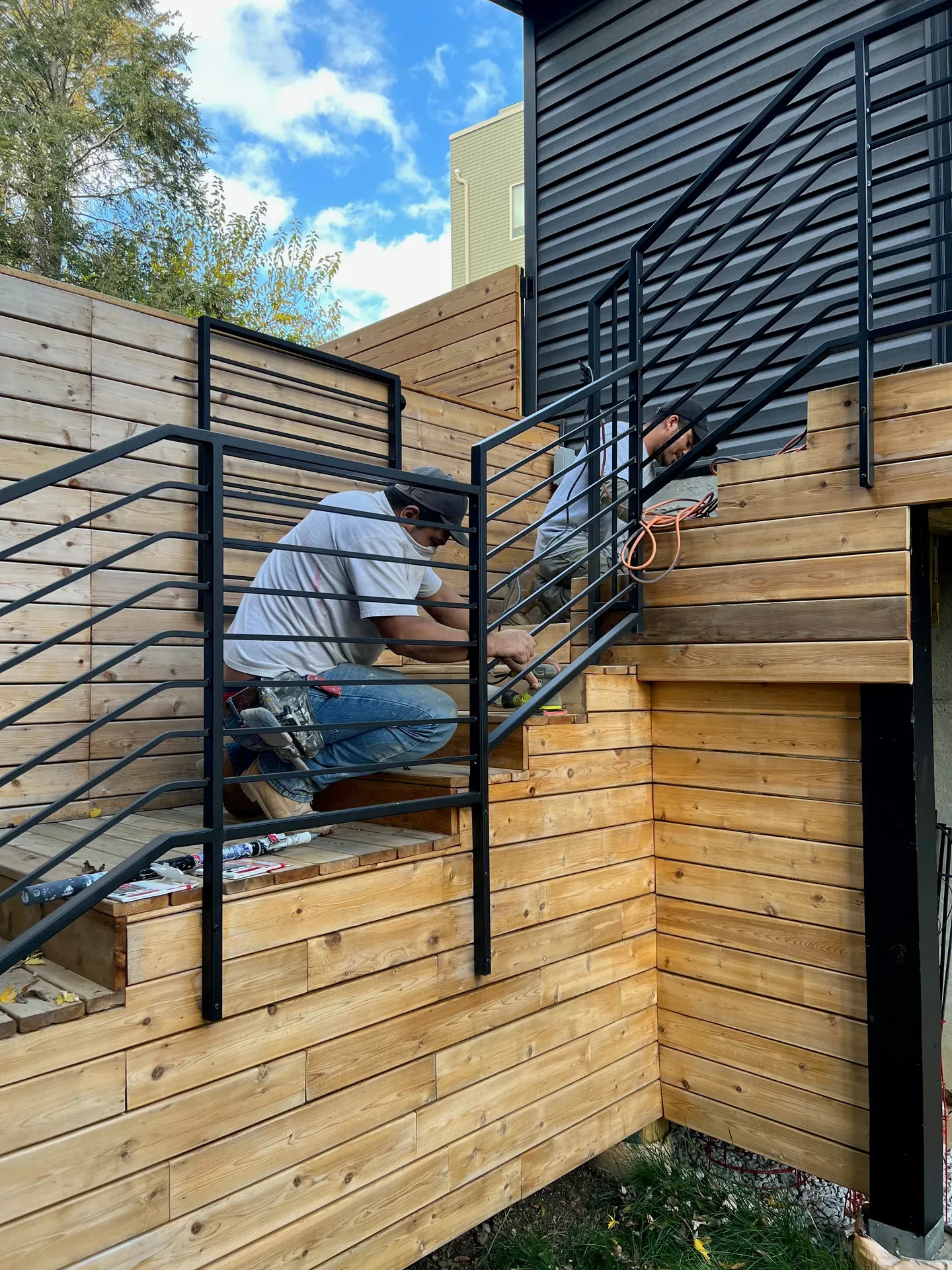 JL crew member kneeling on a freshly-built cedar tongue-and-groove exterior stair landing, fabricating a black powder-coated steel railing connection, urban rowhouse setting