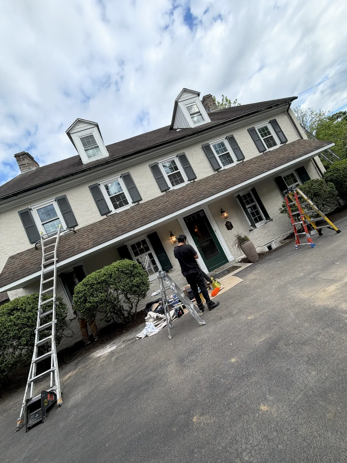 Large white Pennsylvania colonial mid-repaint, JL crew member at the front door working off a step ladder, full-height extension ladders set against the eaves on both ends of the house