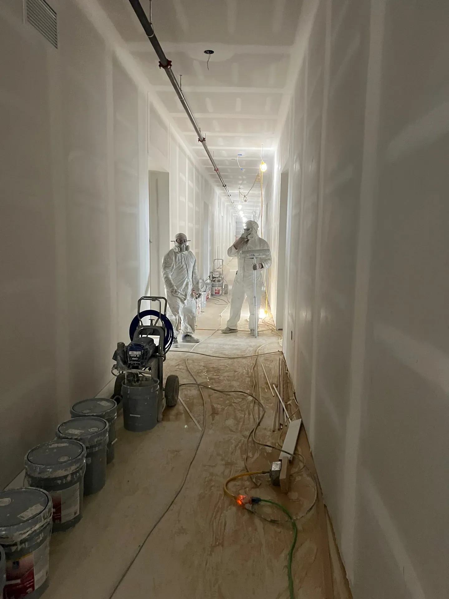Two JL crew members in white Tyvek suits and respirators with an airless spray rig in a long commercial corridor mid-drywall finish, fresh white walls under construction lighting