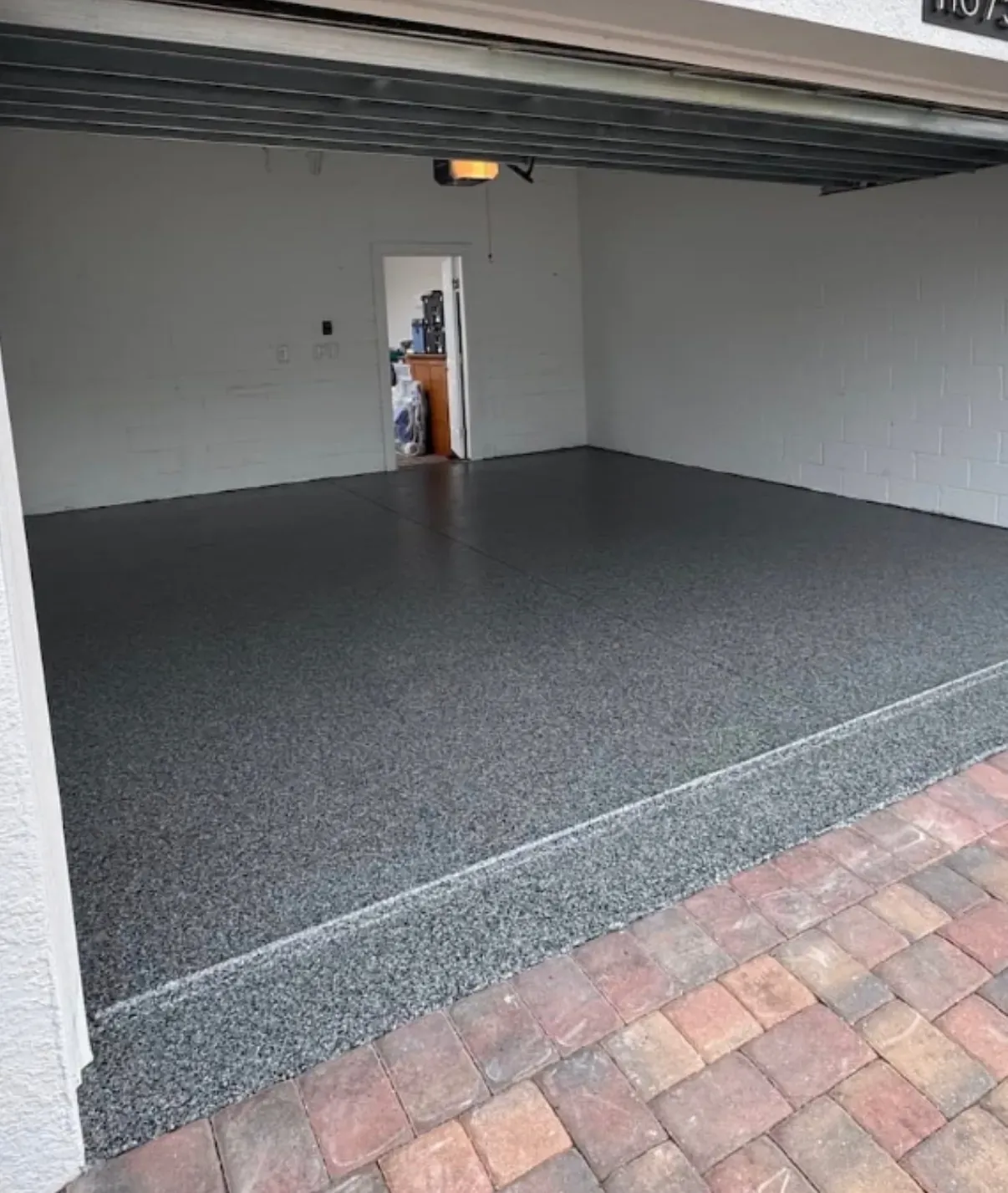 Two-car residential garage with freshly applied gray epoxy and color-flake floor coating, paver apron visible at the door threshold, garage door open and white concrete-block walls visible