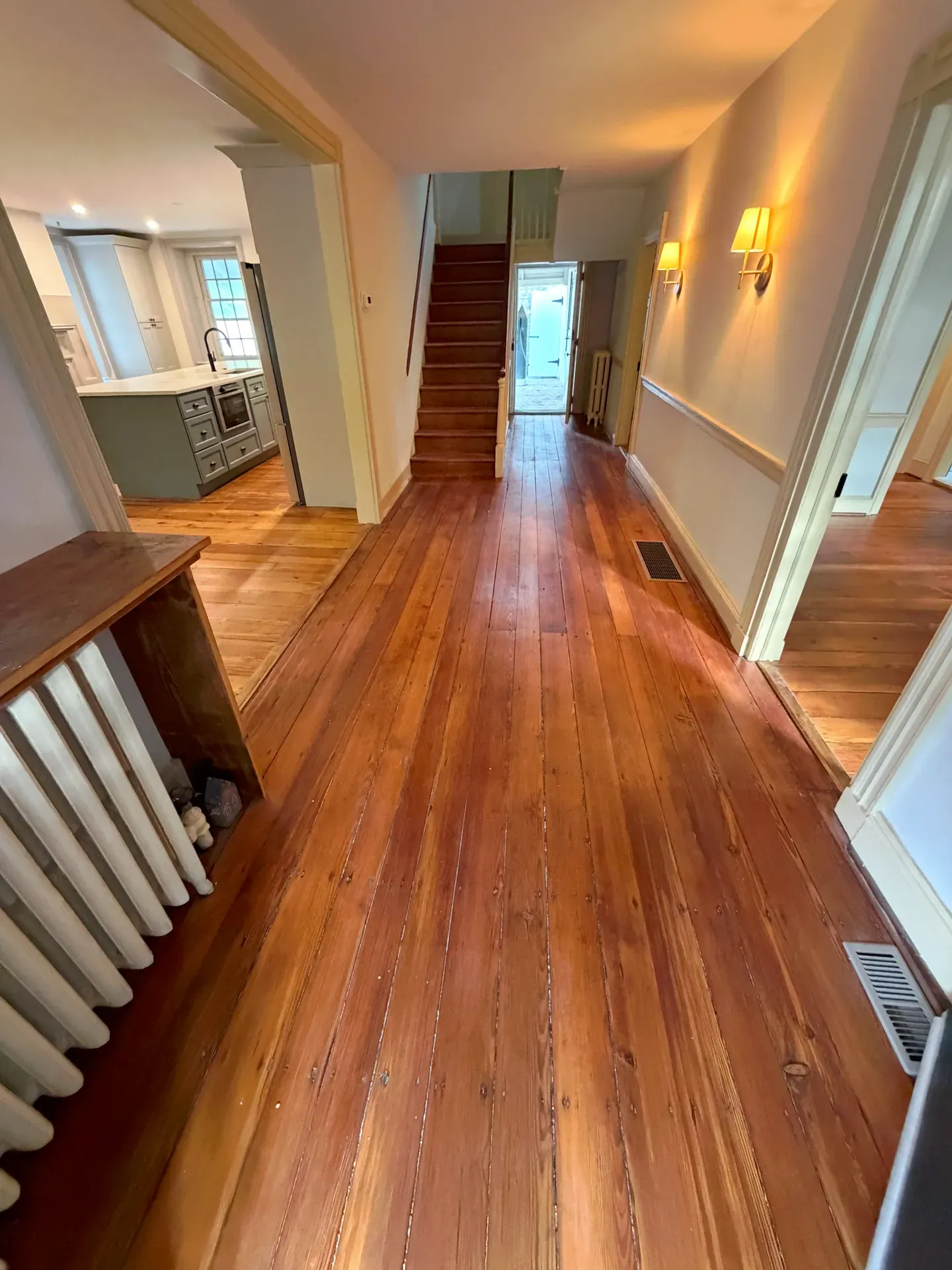 Refinished wide-plank pine floor running down a center-hall in a restored Pennsylvania colonial, view through to a kitchen and dining beyond, original staircase with wood railing on the left