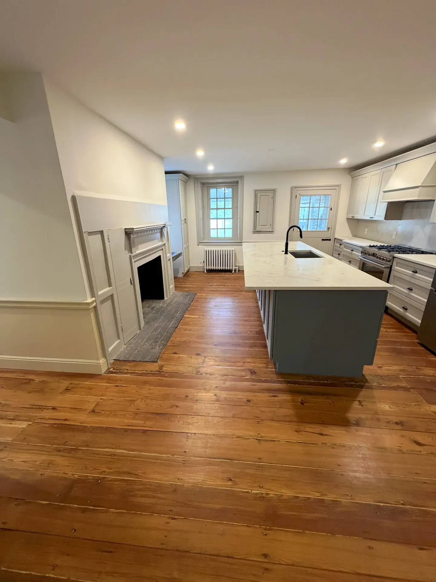 Modern open-plan kitchen with freshly refinished wide-plank pine floors, white shaker cabinets, fireplace mantel, large island with quartz countertop — refinished floor extending into the dining and living spaces