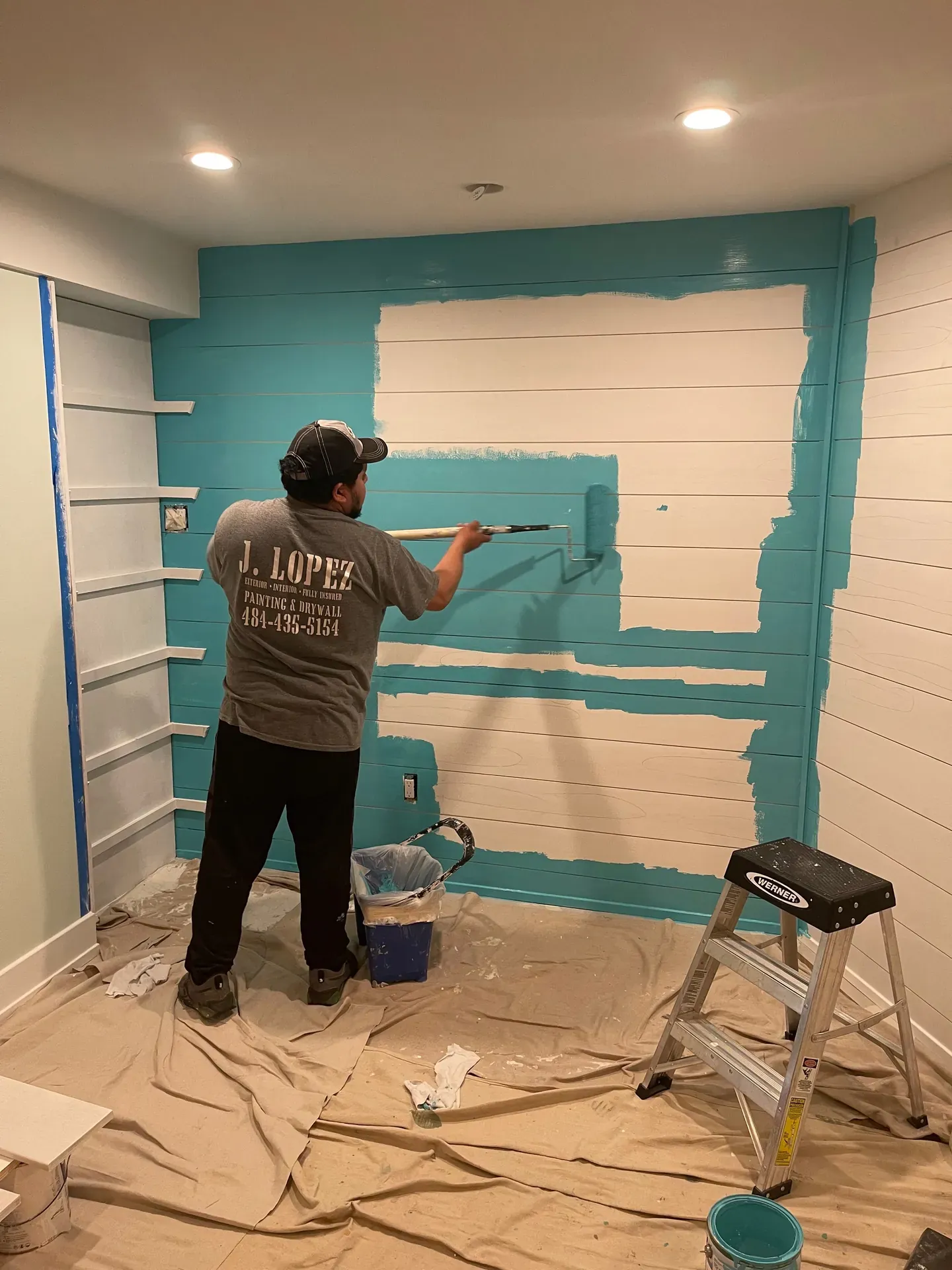 JL crew member in branded J. Lopez Painting & Drywall T-shirt with 484-435-5154 printed across the back, rolling a teal accent color over a white shiplap wall in a finished basement, white shelving on the left, drop cloths on the floor
