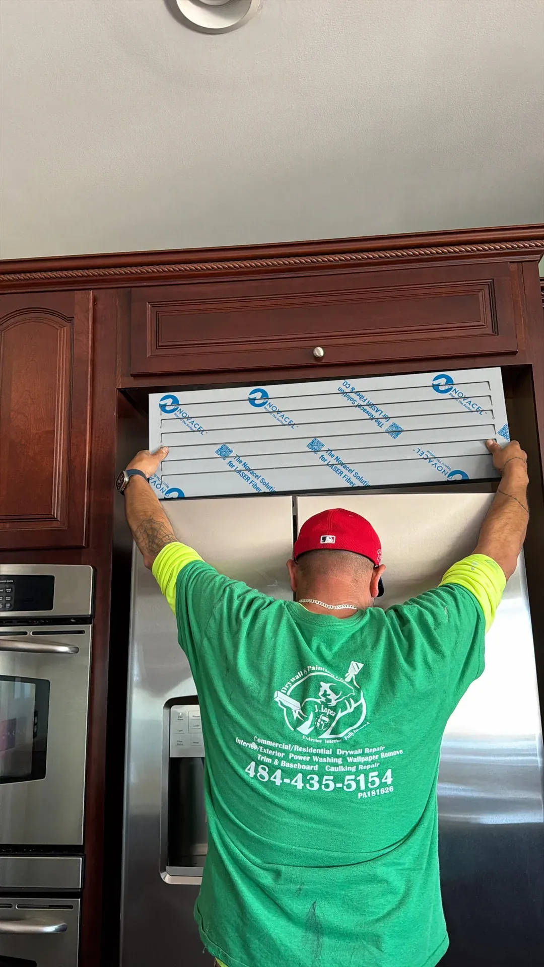 JL crew member in green JL Drywall and Painting branded uniform installing a range hood vent panel between upper kitchen cabinets, dark wood cabinetry and stainless appliances visible