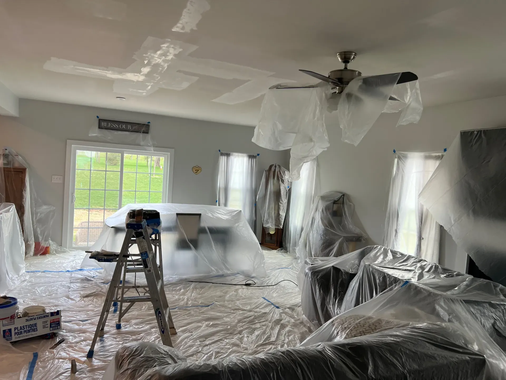 Living room mid-ceiling-drywall-repair — white drywall patches visible on the gray ceiling above, the ceiling fan in place, full plastic-sheeting containment wrapped around the furniture and adjacent walls below the work area