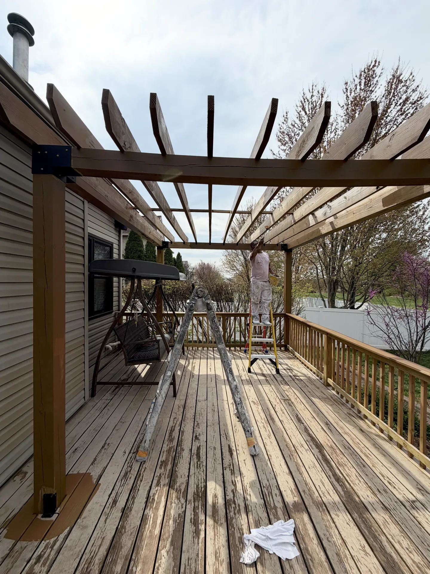 JL crew member on an extension ladder prepping a residential pergola and deck for refinish, weathered cedar surfaces visible, swing chair and ladder in foreground, gray Pennsylvania sky overhead