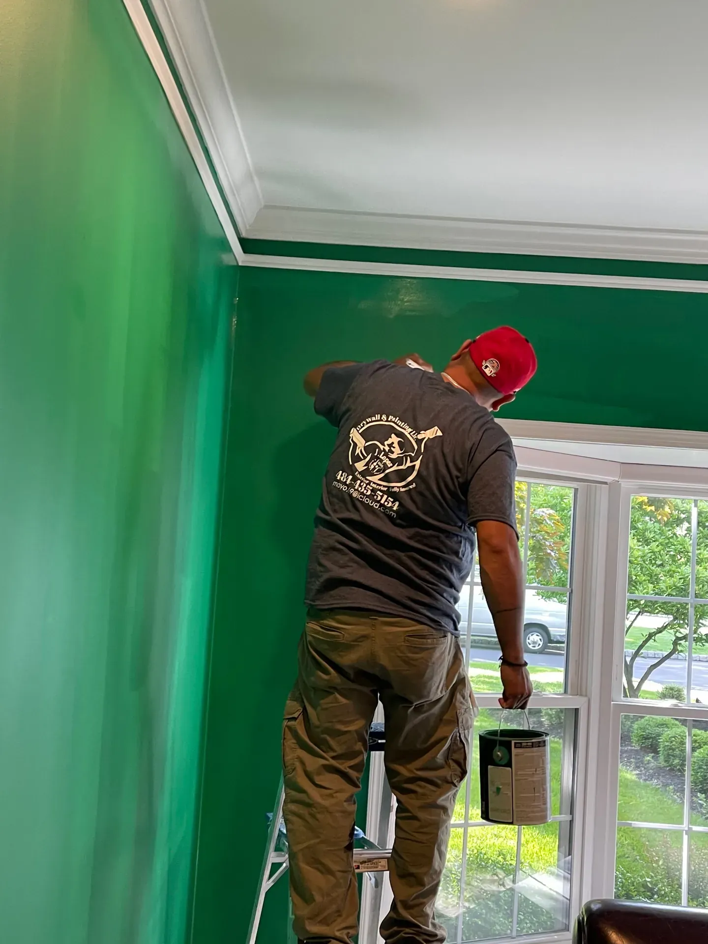 JL crew member in red cap and branded gray T-shirt cutting in a deep emerald-green paint color along the white crown molding of a residential dining room, bay window showing a suburban Pennsylvania street outside
