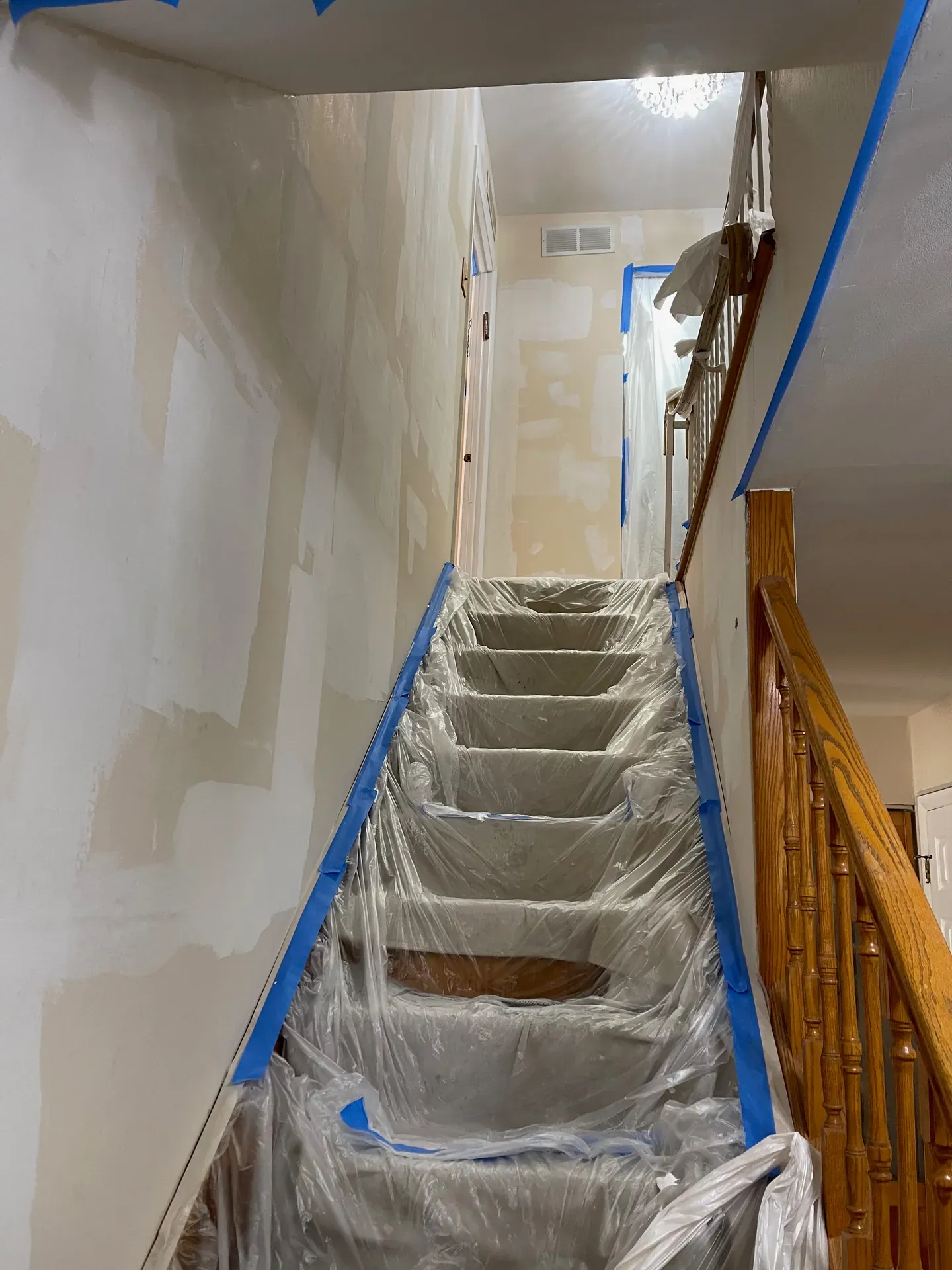 Residential stairwell mid-drywall finish — extensive skim coat work visible on both side walls, blue painters tape masking the railings and trim, plastic sheeting wrapped around every stair tread for full containment, oak railing on the right