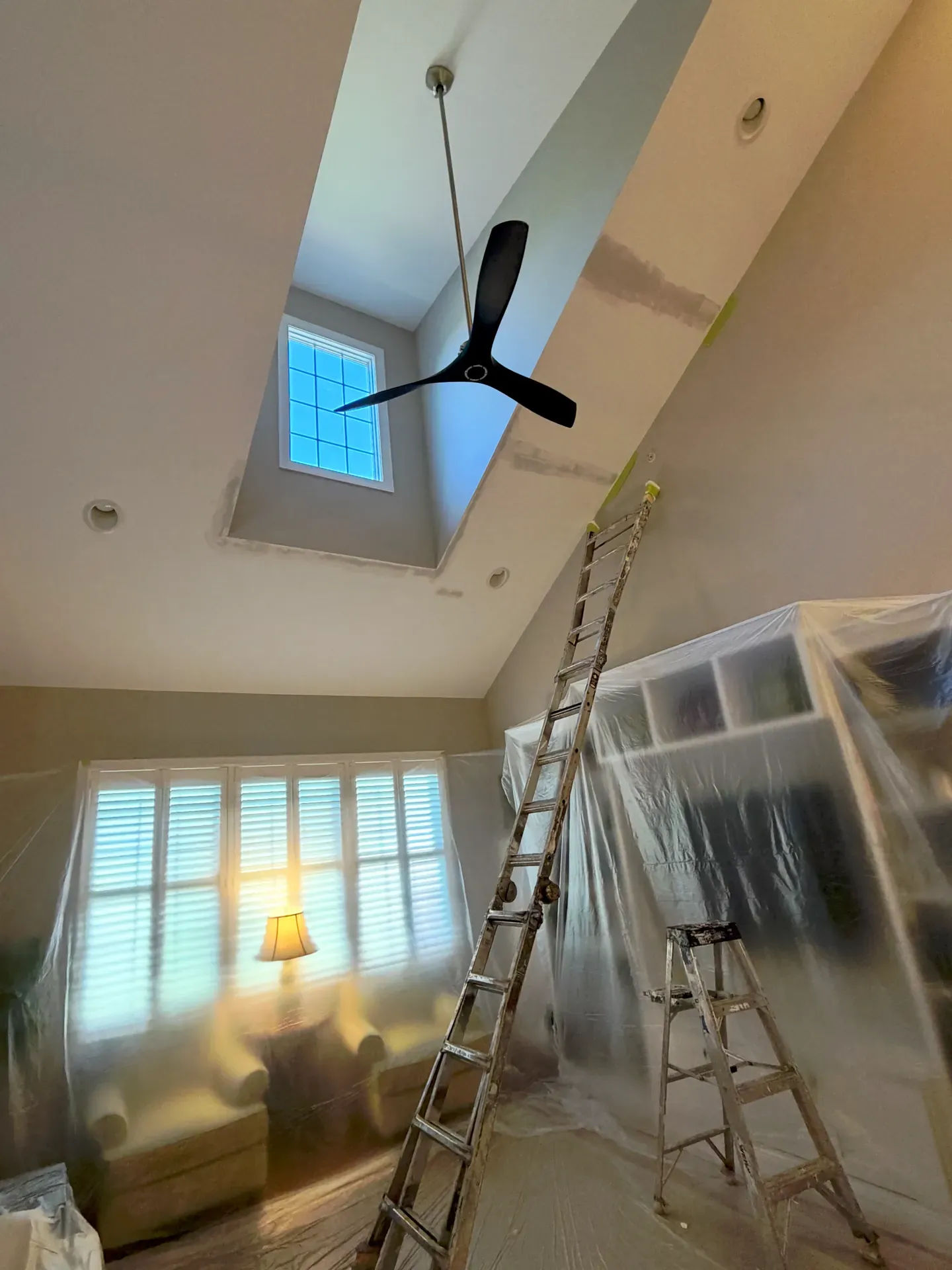 Vaulted residential ceiling mid-paint with the ceiling fan masked, ladder and step ladder set up, plastic sheeting protecting the furniture and shutters below